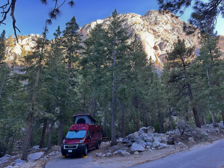 Red RV rental camper van at a wooded campsite, framed by trees against a sunny granite mountain.. Winnebago Winnebago 2023