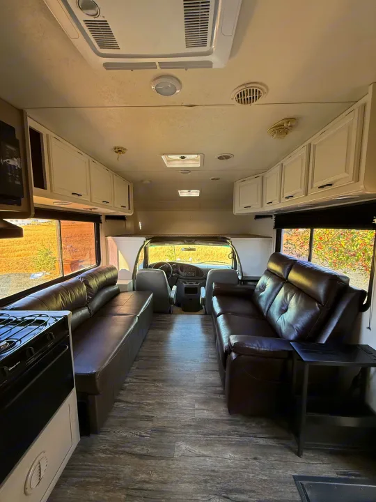 Interior of an RV rental: brown leather sofas, wood-look flooring, white cabinets, and the driver's cockpit view.. Coachmen Leprechaun 2002