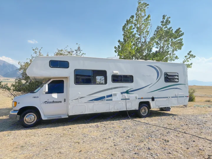 White Coachmen RV rental, a motorhome camper, parked outdoors by trees and mountains under a clear blue sky.. Coachmen Leprechaun 2002
