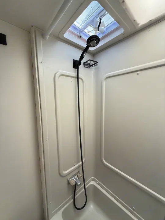 White shower stall inside an RV rental, featuring a black shower head, soap dish, and a skylight with a fan.. Coachmen Leprechaun 2002