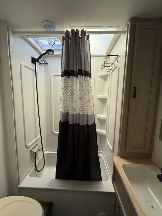 This functional RV rental bathroom features a shower with a patterned curtain, vanity with a sink, and toilet. A skylight brightens the space.. Coachmen Leprechaun 2002