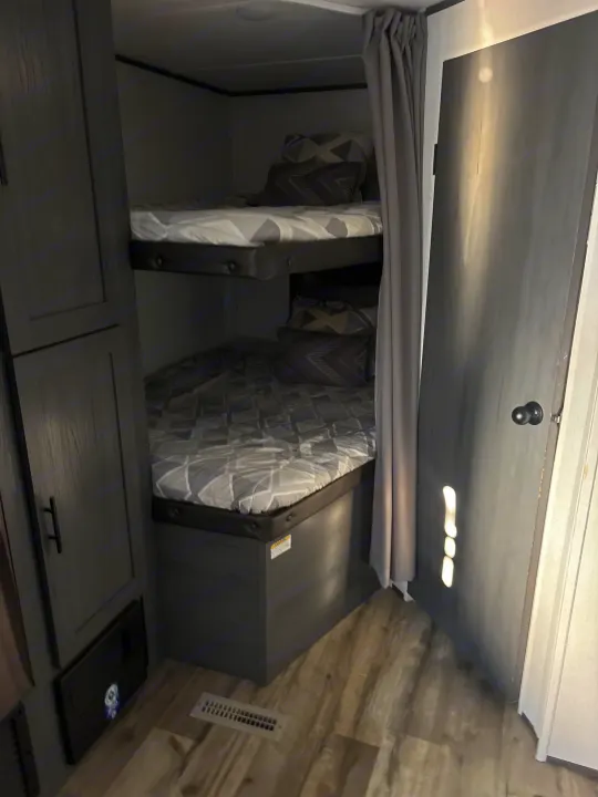 This RV rental features a modern dark grey interior, showcasing two bunk beds on the left and a door on the right.. Dutchmen 26bhc 2024