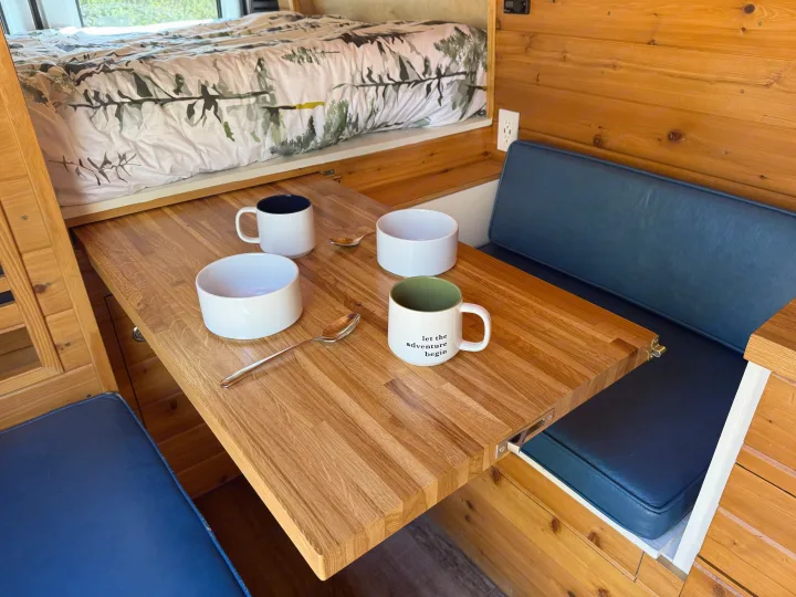 Step inside this inviting RV rental with a built-in wooden table, comfy blue cushions, and a cozy bed. Adventure awaits!. RAM Promaster 2500 2020