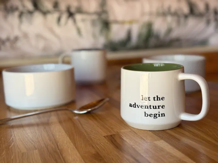A white mug with "let the adventure begin" sits on a wooden table, with other cups and a spoon nearby. Perfect for your RV rental trip!. RAM Promaster 2500 2020