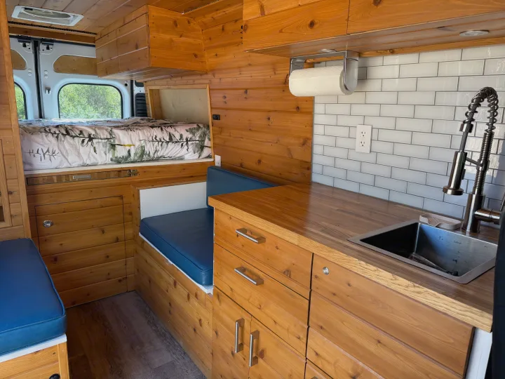 Custom camper van interior, ideal for RV rental. Features wood paneling, kitchen, cozy bed, and dinette with blue cushions.. RAM Promaster 2500 2020