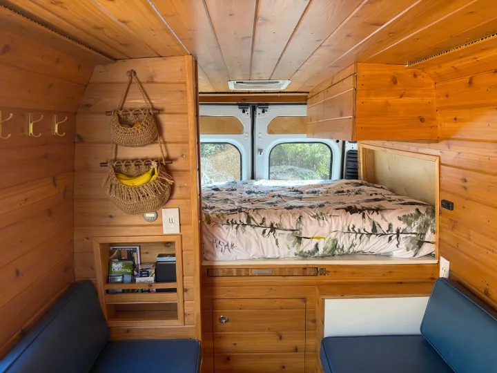 Inside an RV rental: a warm wood interior with a nature-themed bed, clever storage, and comfy blue seats.. RAM Promaster 2500 2020