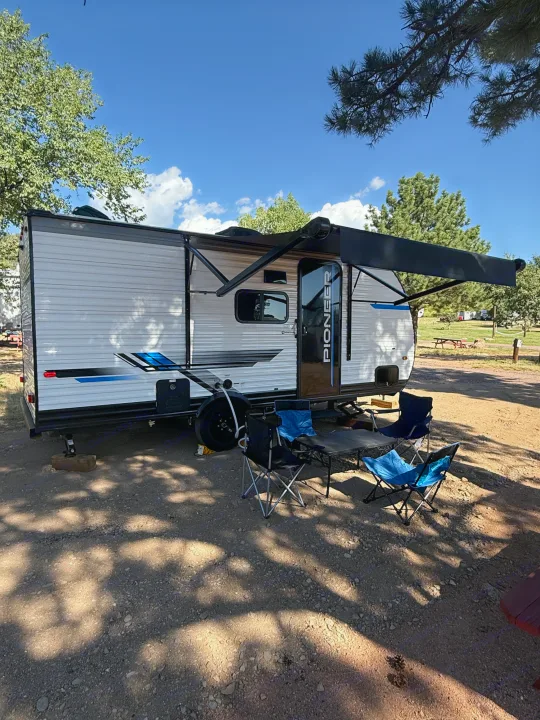 A modern RV rental with its awning extended, accompanied by camping chairs and a table on a shaded campsite.. Heartland RVs Pioneer 2025
