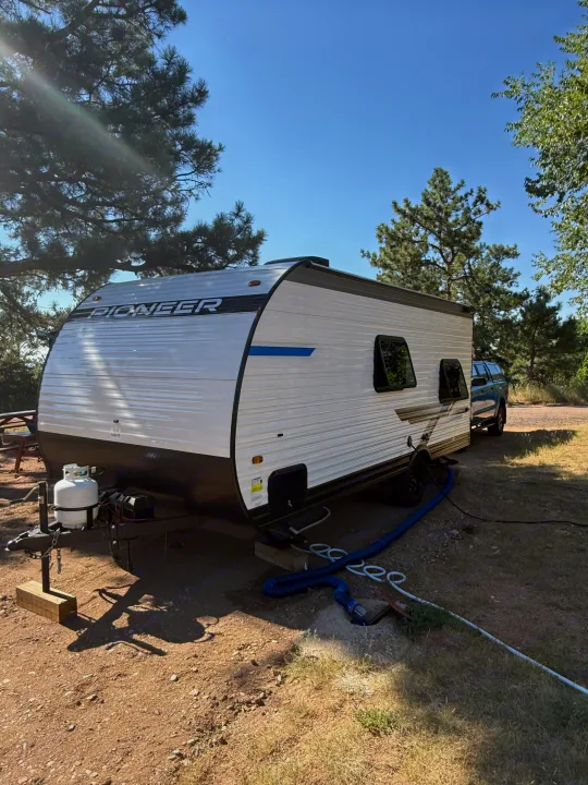 A white Pioneer RV rental is parked at a sunny campsite with a propane tank, utility hookups, trees, and blue sky.. Heartland RVs Pioneer 2025