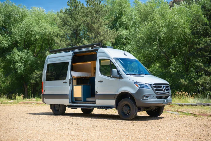 Spacious light gray camper van, perfect for rv rental trips. Open door reveals cozy interior, parked on gravel by trees.. Geotrek Flatiron Summit 2025