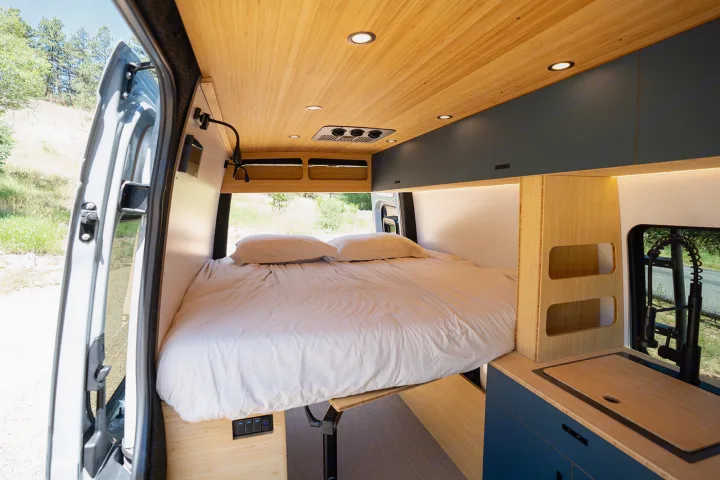 Cozy rv rental van interior: a large bed, sleek bamboo ceiling with lights, and a compact kitchen. Nature visible outside.. Geotrek Flatiron Summit 2025