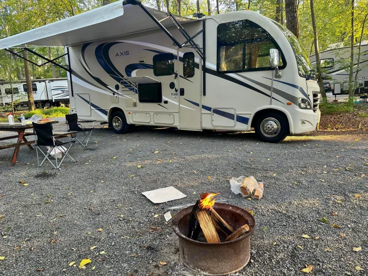 An RV rental parked at a serene campsite with an extended awning, picnic table, chairs, and a burning campfire.. Thor Axis 2018