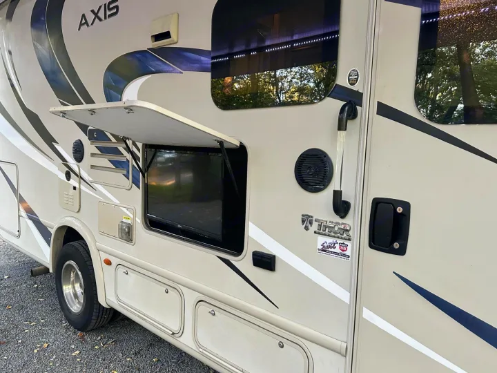 This RV rental features an outdoor entertainment setup with a TV and speaker, plus exterior storage, perfect for camping.. Thor Axis 2018