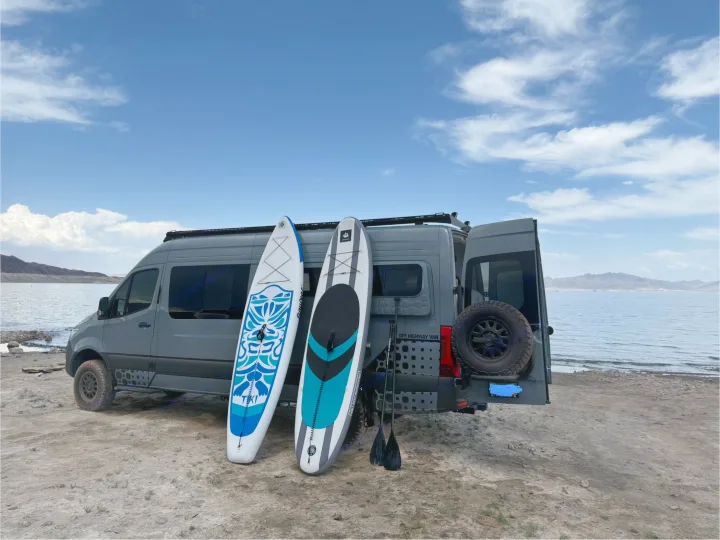 This grey rv rental adventure van with paddleboards is parked lakeside, ready for water sports fun. Ideal for outdoor exploration.. Off Highway Van Off Highway Van Class B 2024