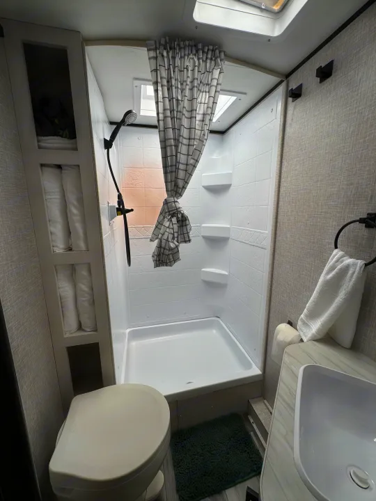 This RV rental bathroom features a compact shower, toilet, sink, and built-in shelving with neatly folded towels.. Jayco Jay Flight 197MB 2025