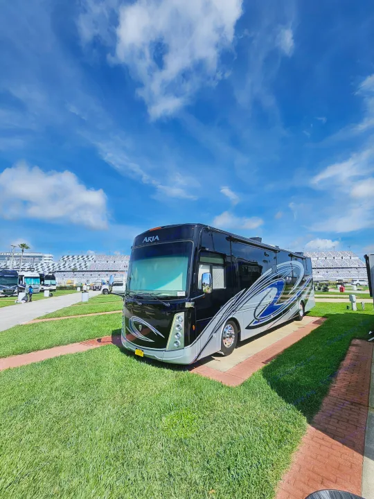 A luxury black and silver Aria RV rental sits on green grass with brick accents, overlooking a race track grandstand.. Thor Motor Coach 2023