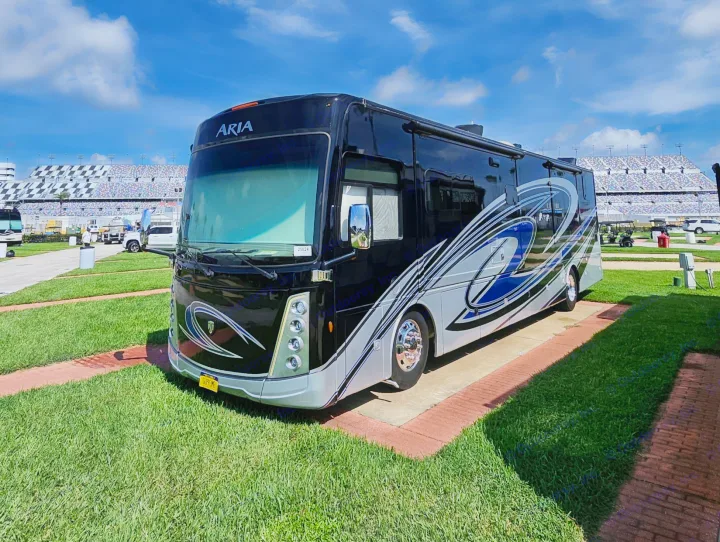 Black and silver Aria RV parked at a stadium, ideal for an RV rental. The sunny scene features green grass and a large grandstand.. Thor Motor Coach 2023