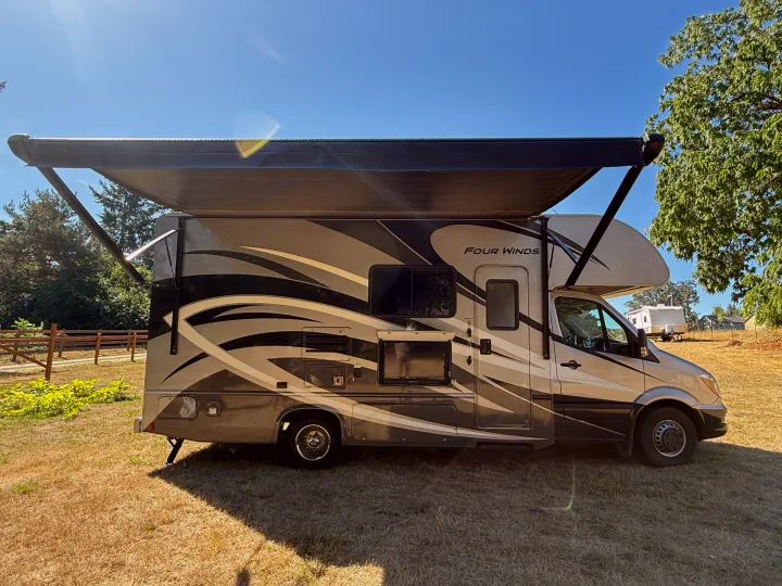 A Class C motorhome with an extended awning, ready for an RV rental trip, is parked on dry grass under a clear blue sky.. Thor Four Winds 2019