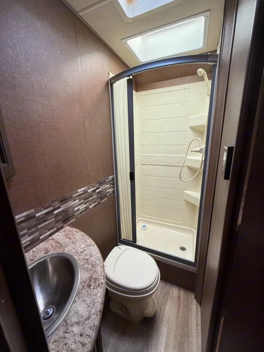 Clean RV rental bathroom featuring a compact toilet, corner sink, and white shower enclosure with textured brown walls.. Thor Four Winds 2019