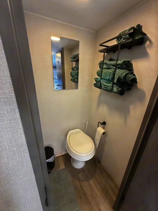 Small RV rental bathroom with white toilet, mirror reflecting the interior, and green towels on wall shelves.. Heartland RVs Pioneer 2025