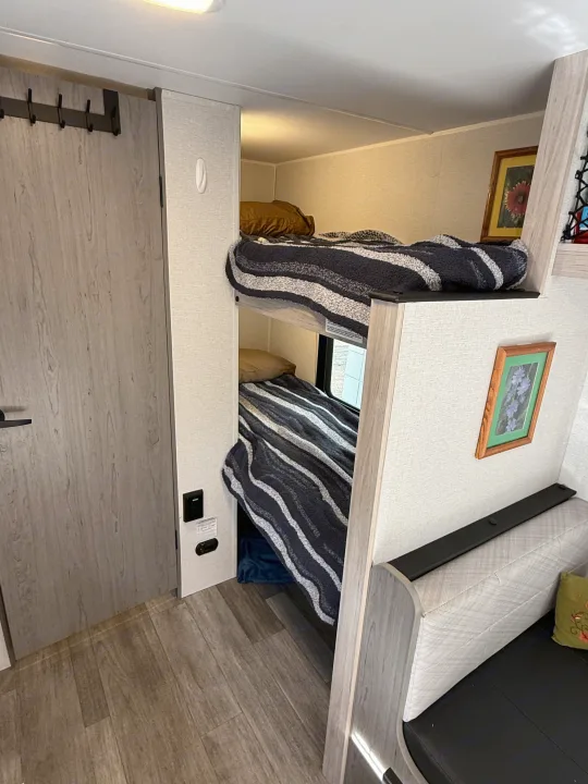 Compact RV rental interior featuring stacked bunk beds with striped bedding, a small dinette, and wood-look floors. Ideal for travelers.. Heartland RVs Pioneer 2025