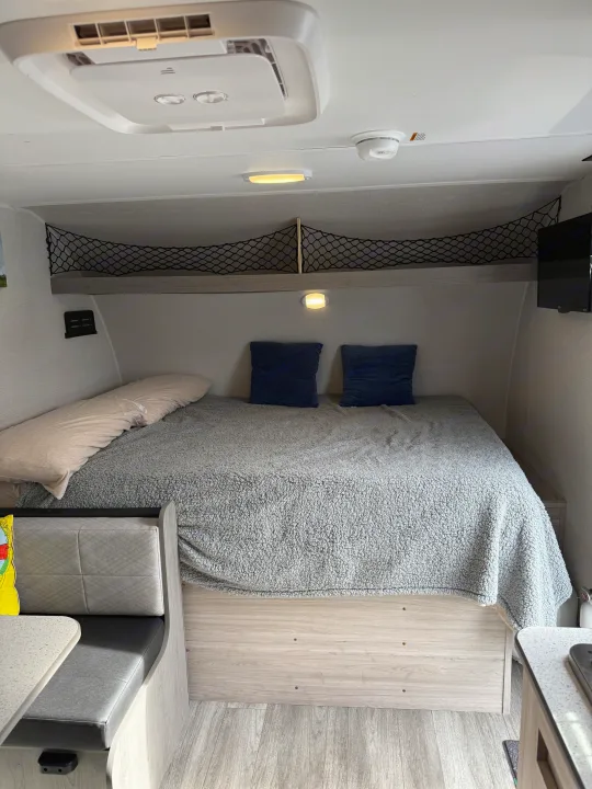 Cozy RV rental interior featuring a comfortable bed with grey blanket, overhead storage net, and dinette area.. Heartland RVs Pioneer 2025