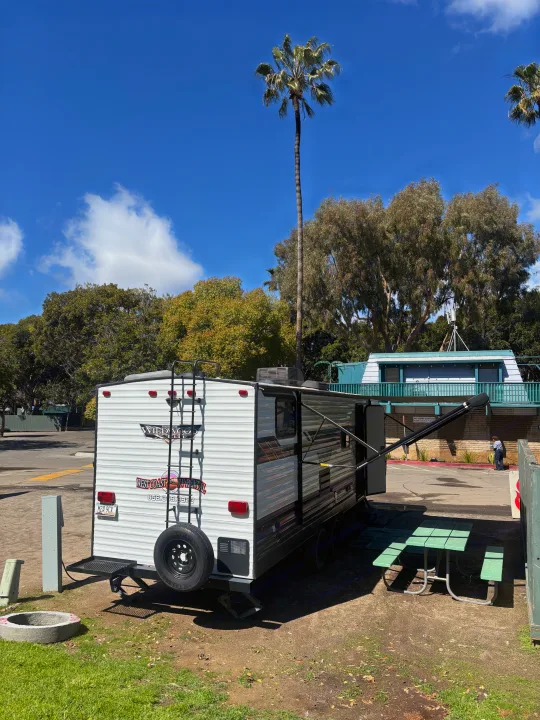An RV rental travel trailer is parked at a sunny campsite with a picnic table, fire pit, and tall palm trees.. Forest River Wildwood X-Lite 2023