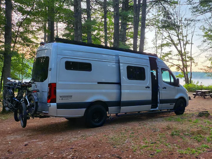 Silver camper van RV rental parked by a misty lake amidst trees, bikes on back. Ideal for nature camping adventures.. Winnebago Adventure Wagon 2023