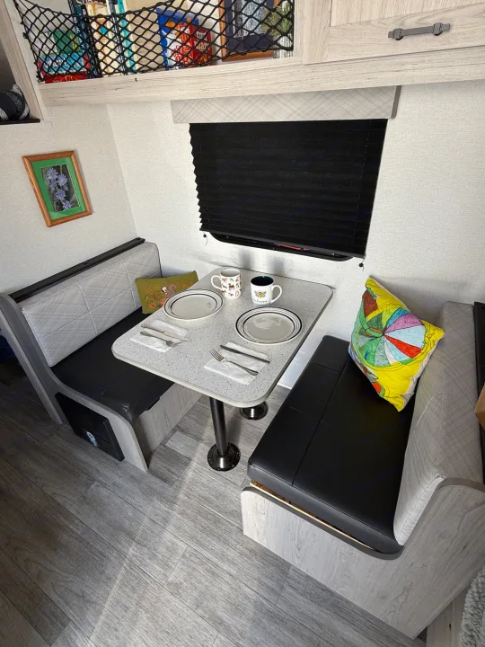 A bright RV rental dinette with two cushioned benches, a central table, and overhead netted storage, set for a cozy meal.. Heartland RVs Pioneer 2025