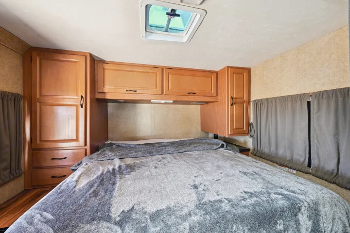 RV rental bedroom featuring a bed with gray blanket, wooden cabinets, overhead storage, side windows with curtains, and a roof vent.. Thor Majestic 2012