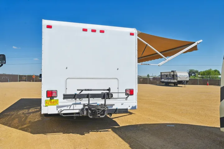 RV rental's rear, with a bike rack and extended awning, parked in a sunny outdoor lot. Another RV is visible in the distance.. Thor Majestic 2012