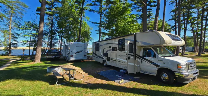 Relaxing at a beautiful rv rental campsite. A Leprechaun Class C RV and another RV sit by a lake with trees and a picnic area.. Coachmen Leprechaun 2021