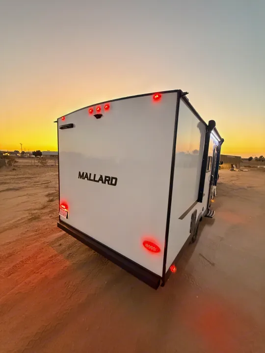 Mallard travel trailer RV rental at sunset, its red lights aglow on a dirt road. Perfect for your next adventure.. Heartland Mallard 2025