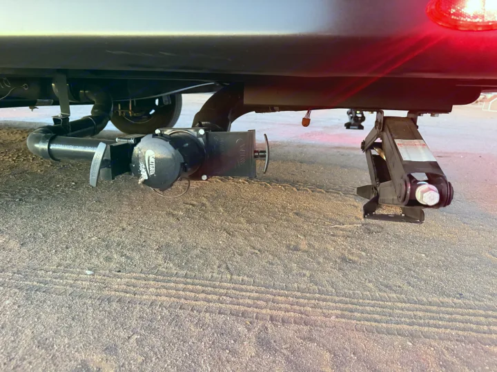 Underside of an RV rental showing its waste water plumbing, dump valve, and a stabilizer jack on sandy ground.. Heartland Mallard 2025