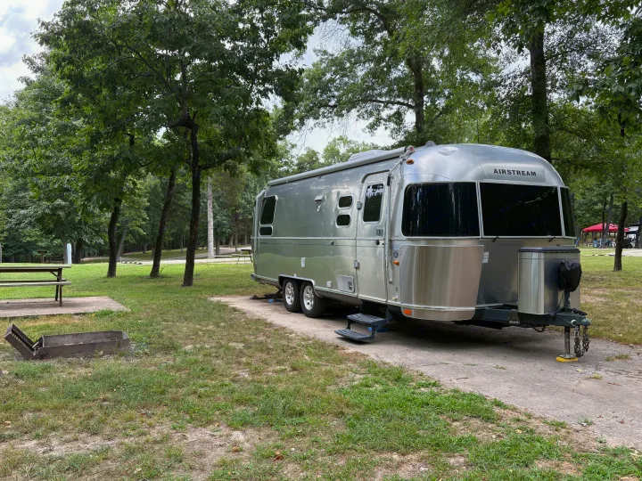 An Airstream RV rental parked at a serene, wooded campsite with a picnic table and fire pit, ready for adventure.. Airstream Flying Cloud 2012