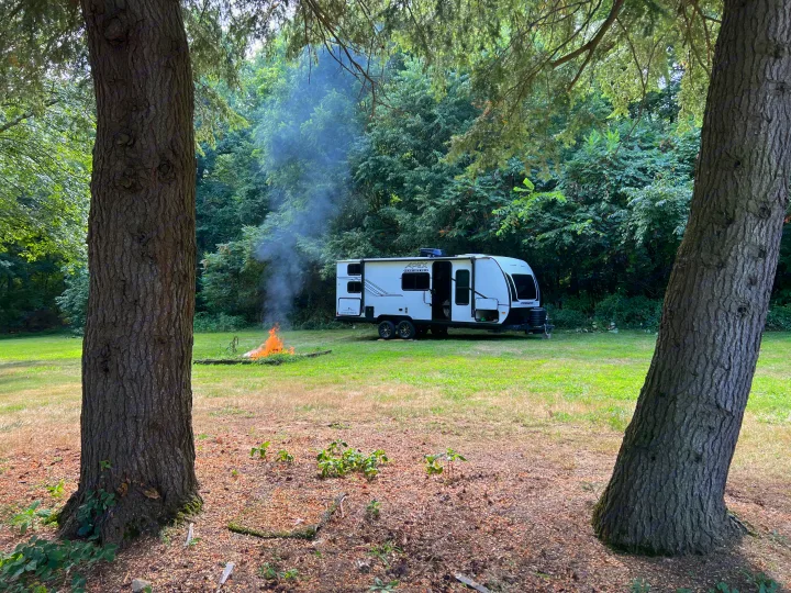 Enjoying a peaceful RV rental camp setup with a warm campfire in a lush forest clearing, framed by two large trees.. Coachmen Apex 2026