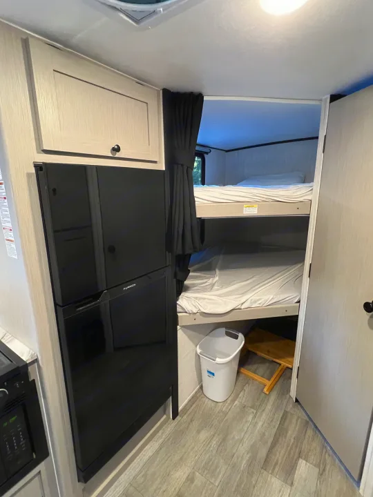 RV rental interior: modern fridge, compact kitchen, and cozy bunk beds with privacy curtain. Perfect for family adventures.. Coachmen Apex 2026