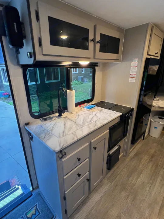Bright RV rental kitchen with modern cabinets, sink, stove, microwave, and an outdoor view.. Coachmen Apex 2026