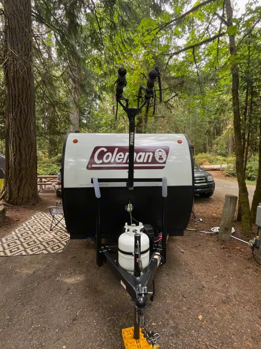 This Coleman RV rental, equipped with a Thule bike rack, is perfectly set up at a wooded campsite, ideal for outdoor adventures.. Dutchmen Coleman Rubicon 2023