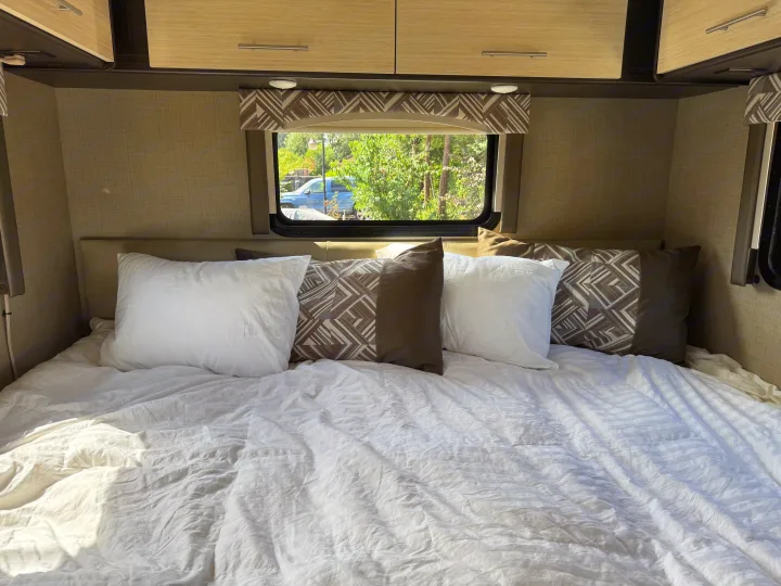 Cozy RV rental bedroom with white bedding, patterned pillows, and a window view of nature.. Thor Axis 2020