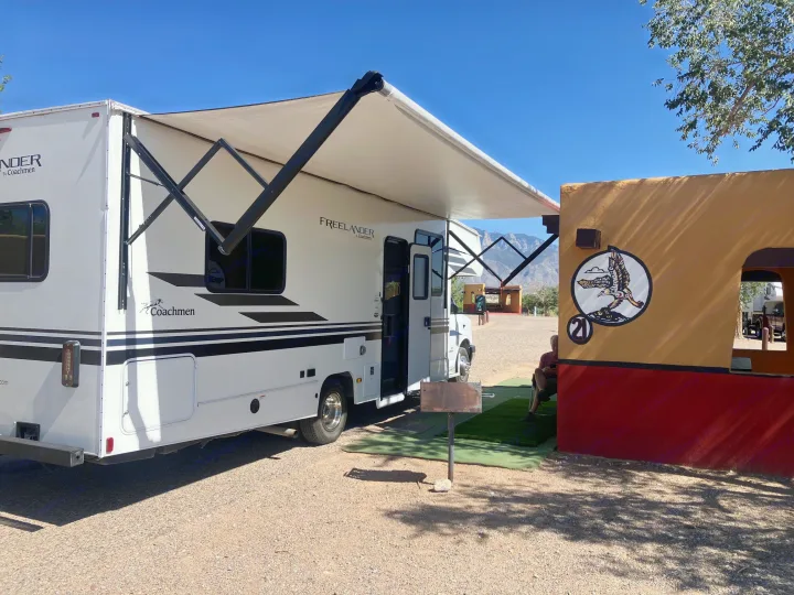 Sunny day for an RV rental: A white Coachmen Freelander RV with an extended awning at a gravel campsite next to a colorful building.. Coachmen Freelander 27QB 2021