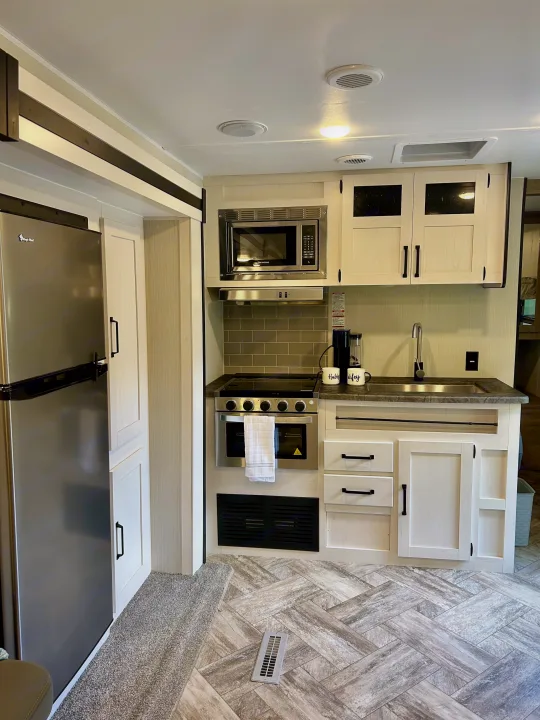 Modern RV rental kitchen featuring stainless steel appliances, light cabinets, sink, and herringbone flooring, offering comfort on the go.. Forest River Palomino Puma XLE 2023