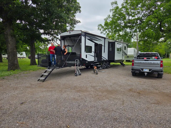 Friends enjoying their RV rental, a toy hauler with a patio deck, at a leafy campground. A pickup truck is parked nearby.. Forest River Vengeance Vengeance Rogue Amored Toy Hauler 2022
