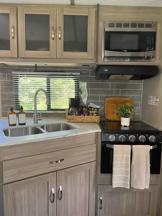 The well-equipped kitchen of an RV rental, showcasing light wood cabinets, a sink, microwave, and stovetop with hanging towels.. Coachmen FREEDOM EXPRESS 2021