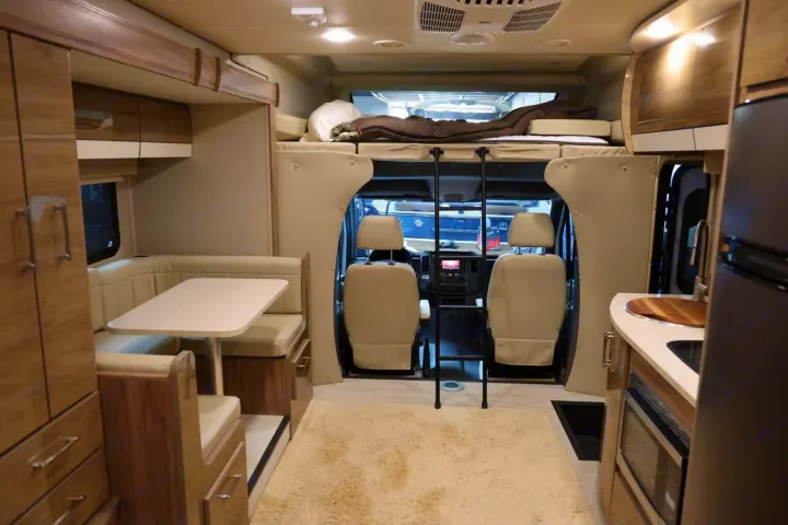 Warm-toned RV rental interior with light wood cabinetry, cream seating, a dinette, kitchen, and overhead bunk.. Entegra Coach Qwest 2019