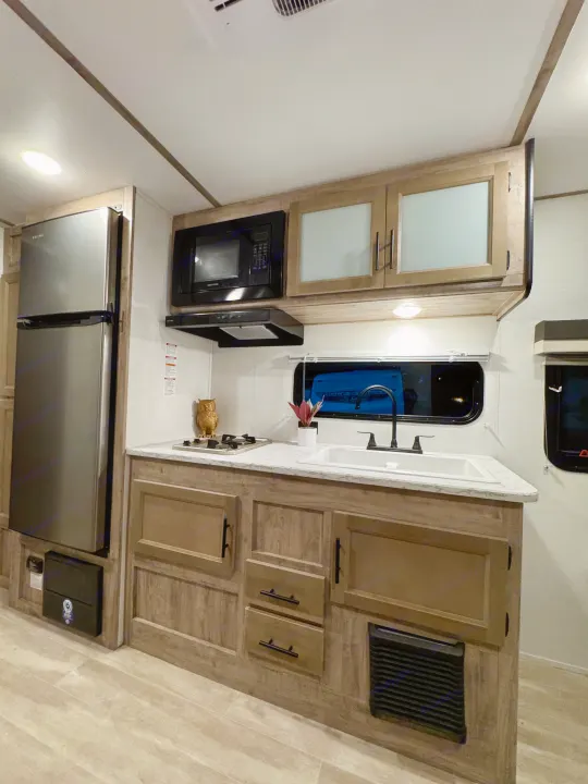 Compact and modern kitchen in an RV rental, featuring a fridge, microwave, sink, stovetop, and wood cabinetry.. Gulf Stream Ameri Lite 2025