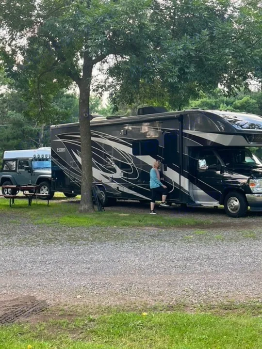 Enjoying the outdoors with an RV rental! A woman stands by a large motorhome at a gravel campsite, with a Jeep parked nearby.. Entegra Coach Esteem 2021
