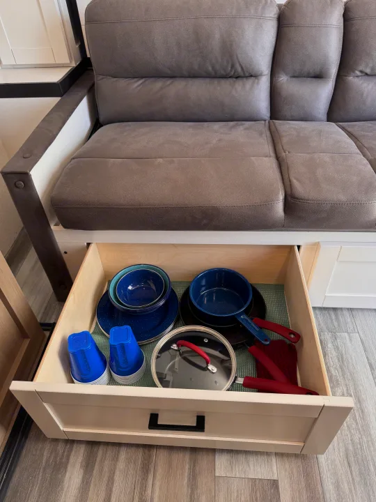 RV rental kitchen: open drawer under sofa showing blue camping dishes, pots, and cups. Efficient travel storage.. Jayco Jay Flight 2026