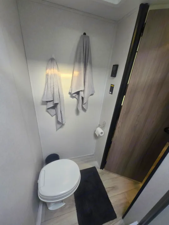 Inside an RV rental bathroom, featuring a compact toilet, hanging towels, and a toilet paper roll. Includes controls & wooden door.. Keystone Colman 2025