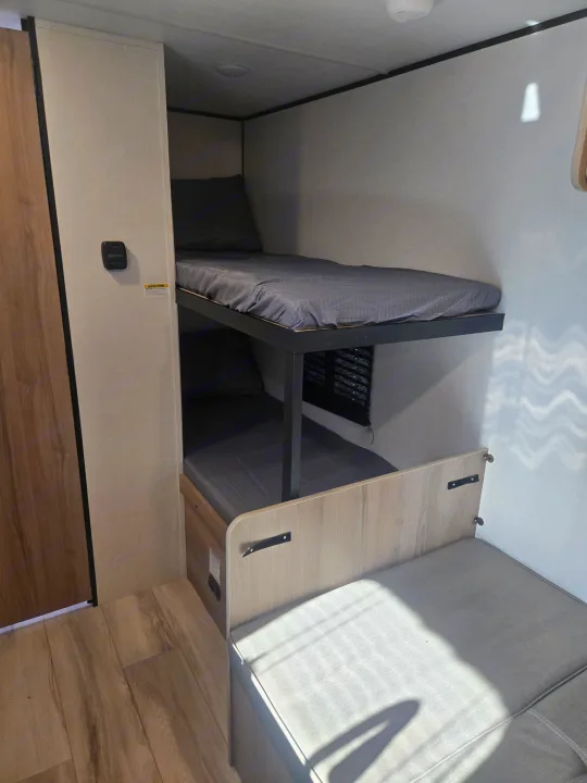 RV rental interior featuring comfortable bunk beds, a wall thermostat, and a light-toned bench.. Keystone Colman 2025