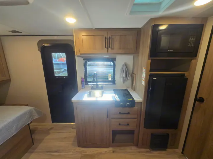 RV rental interior: A compact kitchen with sink, stove, microwave, and fridge, beside a bed and exit door.. Keystone Colman 2025